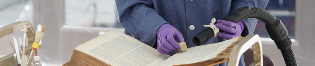 The image shows a conservator, Tiffany Eng Moore, performing a mould remediation treatement on a contaminated book, with a smoke sponge and a vacuum.